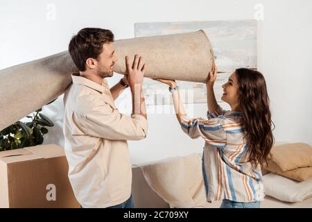 Seitenansicht eines lächelnden Paares, das den Teppich in der Nähe eines Pappkartons im Wohnzimmer hielt Stockfoto