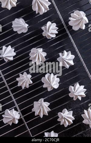 Muster mit einigen weißen selbstgemachten Baiser-Cookies, die auf Stahlrost abkühlen. Strukturierte Tischoberfläche aus schwarzem Stein im Hintergrund. Süßer Hintergrund Stockfoto