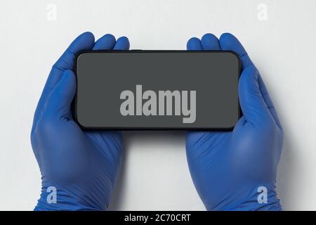 Hände in sterilen medizinischen Handschuhen hält Smartphone auf weißem Hintergrund. Leere schwarze Bildschirmanzeige. Speicherplatz kopieren. Modell. Stockfoto