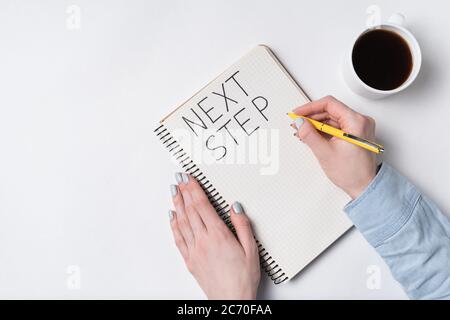 NÄCHSTER SCHRITT Hinweis im Notizblock auf weißem Hintergrund. Handgeschriebene Wörter. Damenhände Tasse Kaffee und Notizbuch. Stockfoto