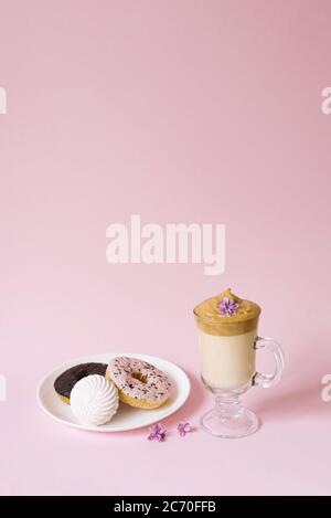 Schöner Dalgon Kaffee trinken. Eine Tasse Kaffee mit Schaum und Fliederblüte, ein Teller mit Süßigkeiten: Marshmallows und Donuts auf einem rosa Hintergrund. Stockfoto