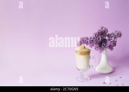Schöne dalgona trinken einen schaumigen Kaffee in einem transparenten Becher und Blumen lila Il lila Hintergrund. Neben dem Becher ist ein Zweig aus Flieder Stockfoto