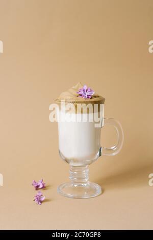 Schöner Dalgon-Kaffeedrink mit Schaum in einer transparenten Tasse und lila Blüten auf beigem Hintergrund. Speicherplatz kopieren Stockfoto