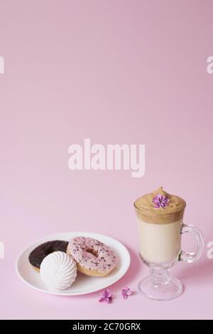 Schöner Dalgon Kaffee trinken. Eine Tasse Kaffee mit Schaum und Fliederblüte, ein Teller mit Süßigkeiten: Marshmallows und Donuts auf einem rosa Hintergrund. Stockfoto