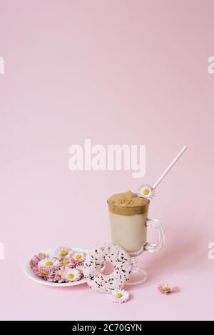 Dalgona Kaffee in einer transparenten Tasse mit Papiertube, einem Teller mit Blumen-Gänseblümchen und einem leckeren leckeren Donat mit rosa Vereisung. Speicherplatz kopieren Stockfoto