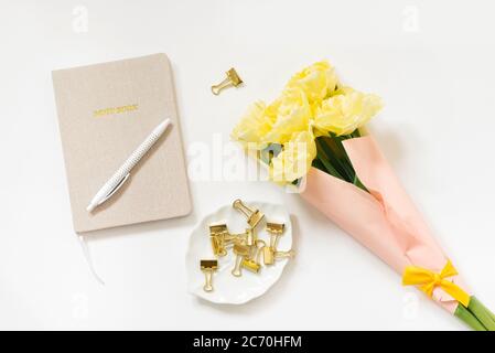 Trending minimalistischen flachen Lay eines Bloggers Arbeitsplatz. Notizblock, Stift, goldfarbene Büroklammern und ein Bündel gelber Tulpen auf weißem Hintergrund. Bis Stockfoto