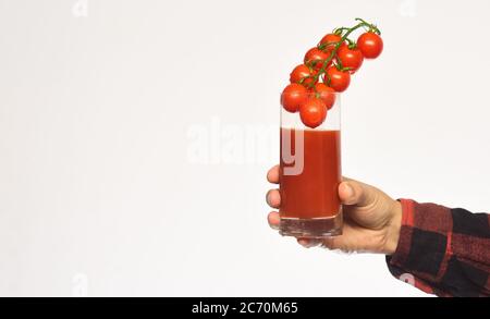 Landwirtschaft und Herbstkonzept. Männliche Hand hält Glas Tomatensaft mit Beeren innen isoliert auf weißem Hintergrund. Mans Handgelenk zeigt rote Kirschtomaten und ihr Produkt. Kerl mit hausgemachten Ernte Stockfoto