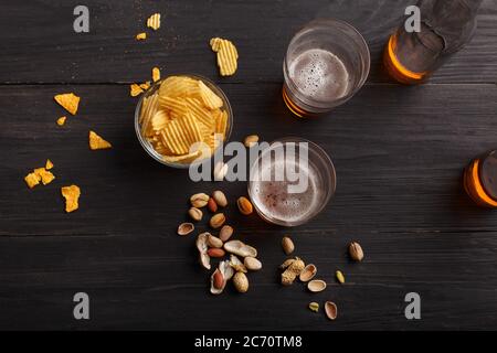 Chips und Pistazien in Glasschale auf dunklem Tisch, daneben zwei Gläser mit hellem Lager und Flaschen Stockfoto