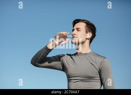 Durst und Sport. Junger Mann in Sportuniform mit modernen kabellosen Kopfhörern, trinkt Wasser Stockfoto