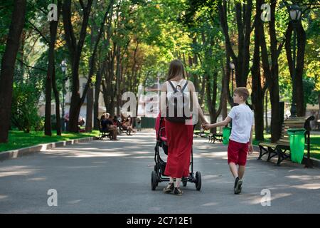 Mama spaziert im Park mit Kinderwagen und älterem Sohn. Gehen Sie mit Kindern. Lifestyle Im Kindesalter. Stockfoto