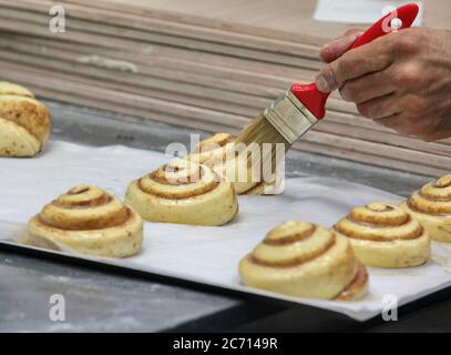 Die geformten Zimtbrötchen dürfen vor dem Backen gesäuert werden, während die Eierverglasung angeraut wird. Fotografiert in einer Boutique-Bäckerei Stockfoto