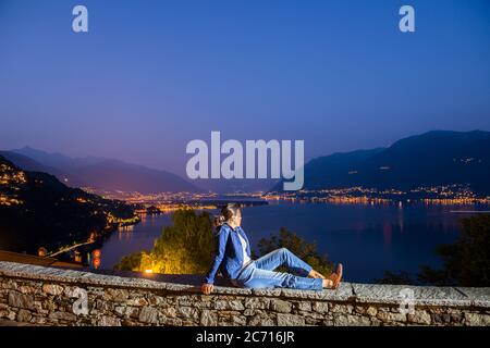 Frau sitzt auf Steinmauer und genießen Panoramablick über den Alpensee Maggiore mit Berg in der Dämmerung in Ronco Sopran Ascona, Schweiz. Stockfoto