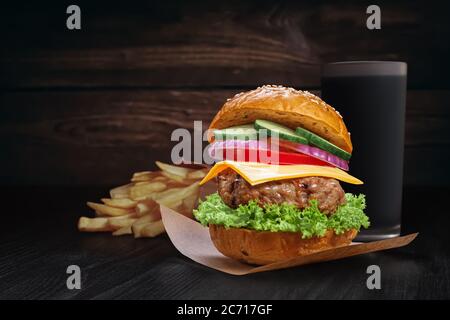 Burger mit Rinderpastete, Käse und Gemüse auf einem Stück Backpapier, Bratkartoffeln und einem Glas Soda auf einem rustikalen Holzhintergrund mit Kopie s Stockfoto