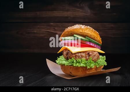 Klassischer Burger aus Brötchen mit Sesamsamen, Rinderpastete, Salat, Käse, Zwiebeln, Gurke, Tomaten auf einem Blatt Backpapier auf einem rustikalen Holzbac Stockfoto