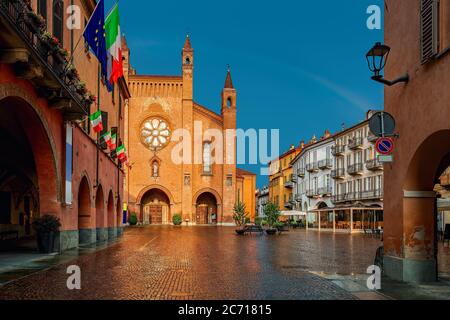 Blick auf den gepflasterten Stadtplatz, alte Häuser und die Kathedrale von San Lorenzo, die von Sonnenlicht unter Regenbogenhimmel in Alba, Piemont, Norditalien beleuchtet wird. Stockfoto