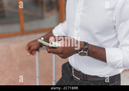 Schwarzer Mann mit Gepäcktasche, der auf der Straße der Stadt mit dem Smartphone steht und formelle Kleidung trägt Stockfoto