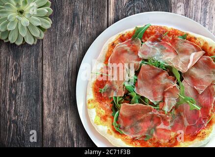 Blick von oben auf frische hausgemachte leckere italienische Pizza mit Serrano Schinken und Rucola auf rustikalem Holztisch Stockfoto