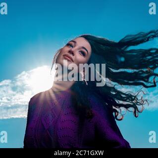 Glückliche Frau mit Locken flattern im Wind Haar auf Blue Bright Sky Hintergrund. Strickpullover oder Pullover in Violett-Strickmuster aus sommerlicher Baumwolle Stockfoto