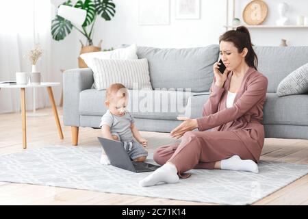 Verärgerte Frau, die auf dem Handy spricht, während ihr Kleinkind Baby den Laptop der Mutter berührt Stockfoto