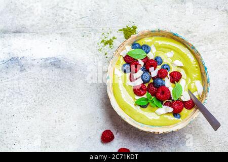 Matcha Tee grüne Smoothie Schüssel mit Beeren und Kokosnuss, grauer Hintergrund. Gesundes veganes Lebensmittelkonzept. Stockfoto