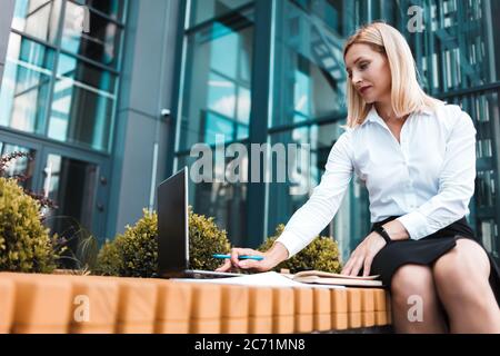 Geschäftsfrau in Bürokleidung sitzt auf einer Bank in der Nähe des Bürogebäudes und nutzt einen Laptop Stockfoto