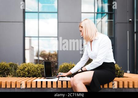 Geschäftsfrau in Bürokleidung sitzt auf einer Bank in der Nähe des Bürogebäudes und nutzt einen Laptop Stockfoto