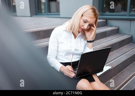 Müde Büroangestellte sitzt auf der Treppe hinter einem Laptop und hält ihren Kopf. Überarbeitungskonzept Stockfoto