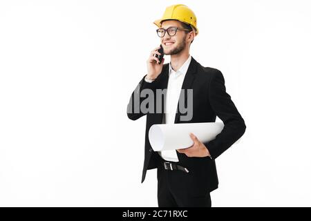 Junger lächelnder Bussinesman in Brillen und gelbem Schutzhelm, der fröhlich zur Seite schaut, während er auf dem Handy mit Plan in der Hand über Weiß redet Stockfoto