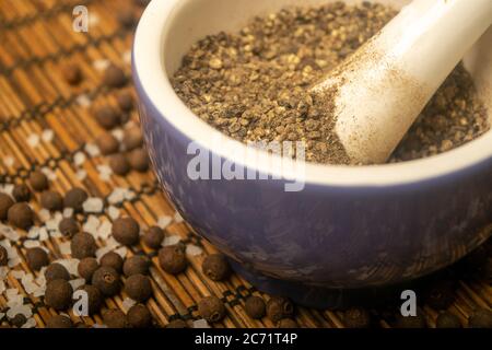 Gemahlenes schwarzes Piment in einem Keramikmörtel und schwarzes Piment und grobes Salz auf einer Schilfmatte verstreut. Nahaufnahme Stockfoto