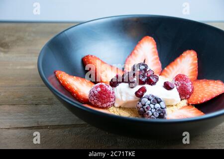 Seitenansicht eines schwarzen Mate Tellers, gefüllt mit geschnittenen Erdbeeren, Keksbrösel, Schlagsahne, Granatapfelkernen, Brombeeren und Erdbeeren auf einem Stockfoto