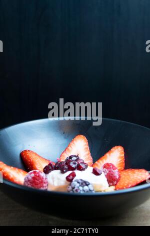 Schwarzer Mate Teller gefüllt mit geschnittenen Erdbeeren, Keks-Krümel, Schlagsahne, Granatapfelkernen, Brombeeren und Erdbeeren auf schwarzem Backgroun Stockfoto