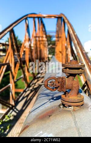 Alte rostige Eisenbrücke über den Fluss mit Dampfleitung. Wärmeversorgung. Industrieanlagen. Detailansicht eines Dampfrohrleitungsventils. Stockfoto