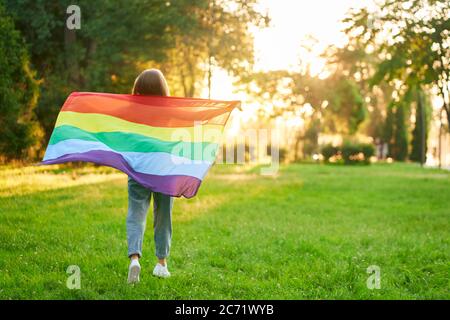 Volle Länge Rückansicht von unkenntlich Mädchen posiert im Park, genießen Sie den Sommer Sonnenuntergang. Inkognito junge Frau mit Regenbogenfahne schwingt auf Wind hinter dem Rücken. Konzept von lgbt, Minoritäten Toleranz. Stockfoto