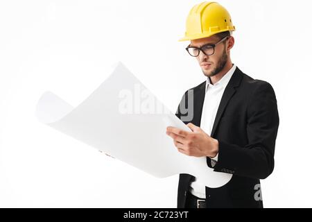 Junge nachdenkliche Bussinesman in Brillen und gelben Schutzhelm stehen mit Plan des neuen Projekts in den Händen über weißem Hintergrund Stockfoto