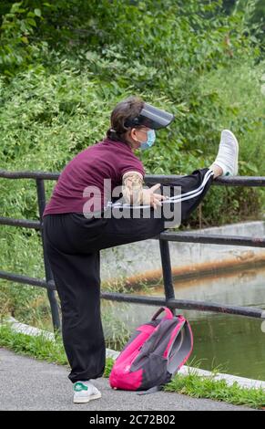 Eine flinke Frau mit einer OP-Maske streckt ihr Bein vor einem morgendlichen Übungsspaziergang. In Kissena Park, Flushing, Queens, New York Stockfoto