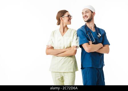 Junge lächelnde Ärztin mit Phonendoskop am Hals und hübscher Krankenschwester in Brillen und Uniform, die sich über weißem Hintergrund fröhlich angucken Stockfoto
