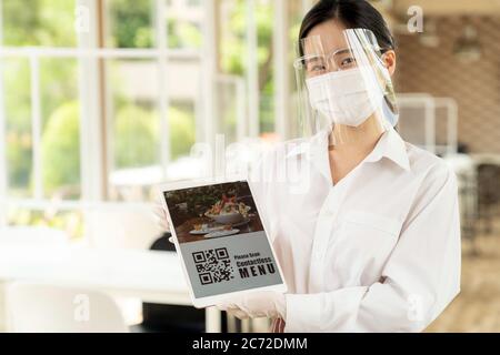 Asiatische Kellnerin mit Gesichtsmaske und Gesichtsschutz halten digitale Tablette mit QR-Code für Kunden für Online-kontaktlos-Menü scannen. Online kontaktlos und Stockfoto
