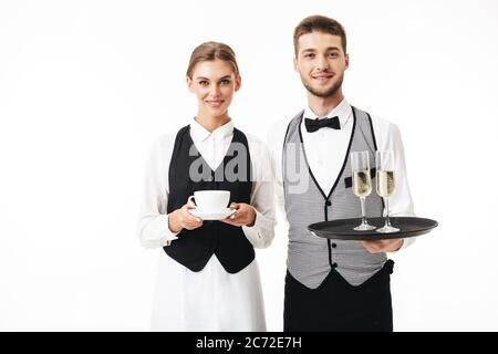 Junge lächelnde Kellner halten Tablett mit Gläsern Champagner, während hübsche Kellnerin halten Tasse Kaffee in den Händen, glücklich in der Kamera zusammen o Stockfoto