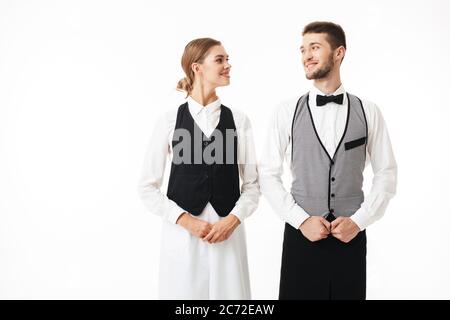 Junge lächelnde Kellner und hübsche Kellnerin in weißen Hemden und Westen glücklich Blick auf einander über weißem Hintergrund Stockfoto