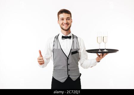 Junge lächelnde Kellner in Uniform glücklich Blick in Kamera halten Tablett mit Brille zeigt Daumen nach oben Geste über weißem Hintergrund Stockfoto