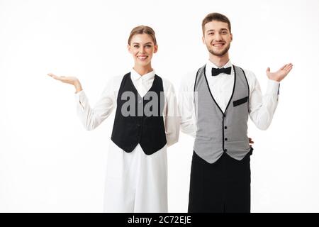 Junge lächelnde Kellner und hübsche Kellnerin in weißen Hemden und Westen glücklich in der Kamera stehend in gleichen Posen über weißem Hintergrund Stockfoto