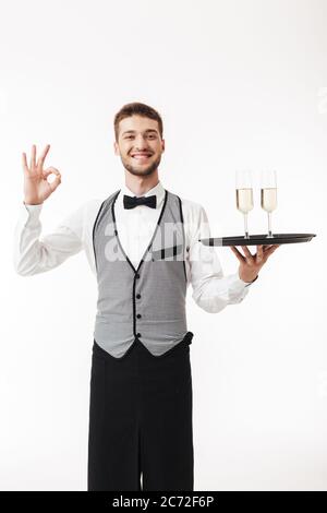 Junger lächelnder Kellner in Uniform, der fröhlich in der Kamera mit Tablett und Brille mit OK Geste auf weißem Hintergrund Stockfoto