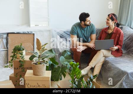 Junges Paar, das sich unterhielt und mit einem Laptop auf dem Sofa in ihrer neuen Wohnung saß Stockfoto