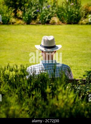 Rückansicht eines älteren Mannes, der im Sommer einen Sonnenhut trägt, auf einer Parkbank sitzt. Stockfoto