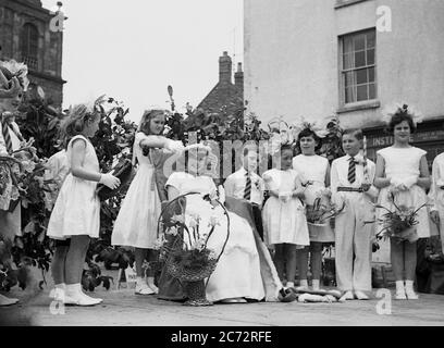 1950er Jahre, historisch, in einer Innenstadt, draußen auf einer hölzernen Plattform, ein junges Mädchen, das auf einem Sitz sitzt und die "May Queen" gekrönt wird, mit anderen kleinen Kindern, die neben ihr stehen, England, Großbritannien. Stockfoto