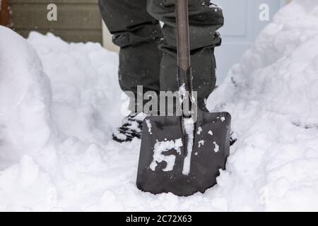 Selektive Fokus und Nahaufnahme auf schwarze Schaufel mit quadratischen Klinge von einem Mann im Hintergrund in Kniehöhe Ansicht gehalten. Schaufeln Schnee nach einem Schneefall. Stockfoto