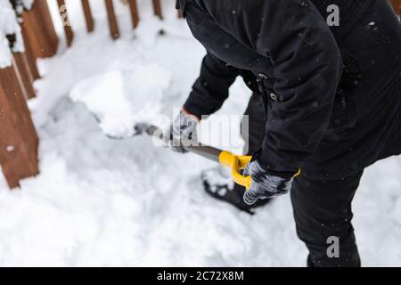 Rückansicht einer nicht erkennbaren Person in Winterkleidung, die sich biegt, während sie die Schaufel voll Schnee hält, schaufelt und nach Schneefall die Hausfront freiräumt. Stockfoto