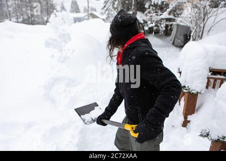 Rückansicht eines Mannes, der den Schnee wirft, um den Gehweg mit einer Schaufel zu räumen, und nach einem starken Schneefall Schnee vom Eingang des Hauses schaufelt. Stockfoto