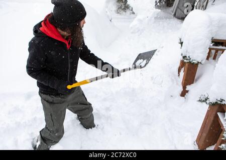 Blick in die Höhe eines jungen Arbeiters, der mit einer Schaufel Schnee wirft, um den Weg zum Hauseingang zu räumen und nach einem starken Schneefall Schnee schaufelt. Stockfoto