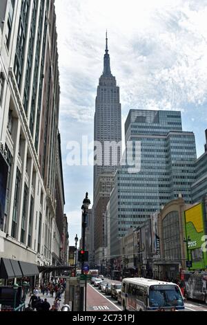 Das Empire State Building, das von einer belebten Straße in New York City aus gesehen wird. Der Verkehr ist entlang der Straße aufgereiht und Fußgänger laufen. Stockfoto